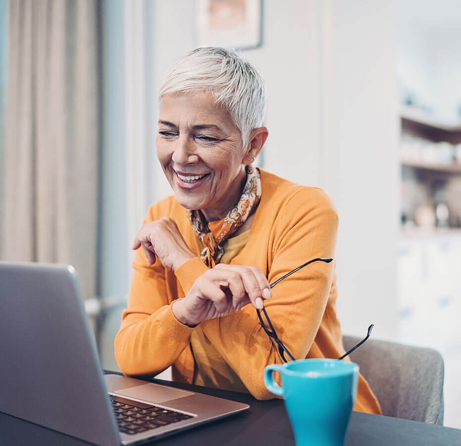 Frau mit kurzem, grauem Haar arbeitet am Laptop, lächelt, trägt Halstuch und hat eine blaue Tasse vor sich.