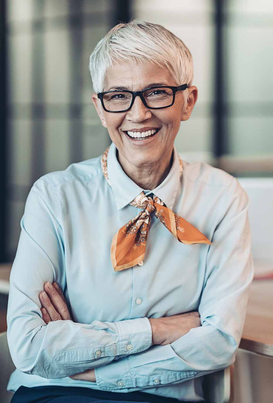 Freundlich lächelnde Frau sitzt mit verschränkten Armen am Tisch. Sie trägt eine hellblaue Bluse und ein orangefarbenes Halstuch. Der Hintergrund ist hell und neutral gehalten.