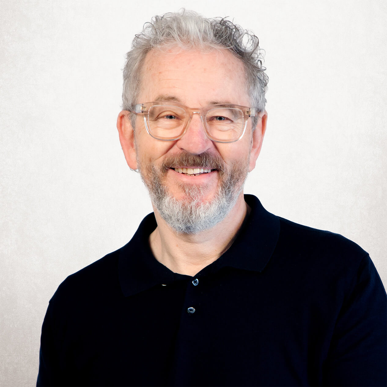 Freundlich lächelnder Mann mit Brille und grauem Bart, bekleidet mit einem schwarzen Polohemd, blickt direkt in die Kamera.